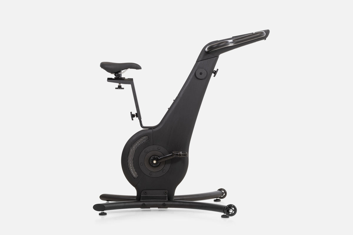 Side view of a black wooden indoor cycle with a black saddle, mesh flywheel cover, and a handlebar touchscreen